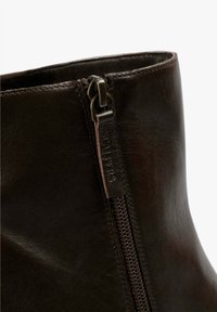Bota de tobillo de cuero marrón que cuenta con una cremallera lateral y una textura suave. La cremallera tiene un tirador de metal oscuro y una lengüeta de cuero con un logotipo en relieve.