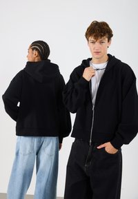Sudadera negra con cremallera y un ajuste relajado, que cuenta con capucha y hombros caídos. Combinada con jeans de pierna ancha azul claro en un modelo y jeans oscuros en el otro.