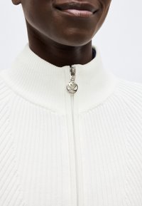 Κοντινό πλάνο ενός ατόμου που φοράει λευκό ριμπ πλεκτό πουλόβερ με ασημένιο φερμουάρ διακοσμημένο με σχέδιο καρδιάς.