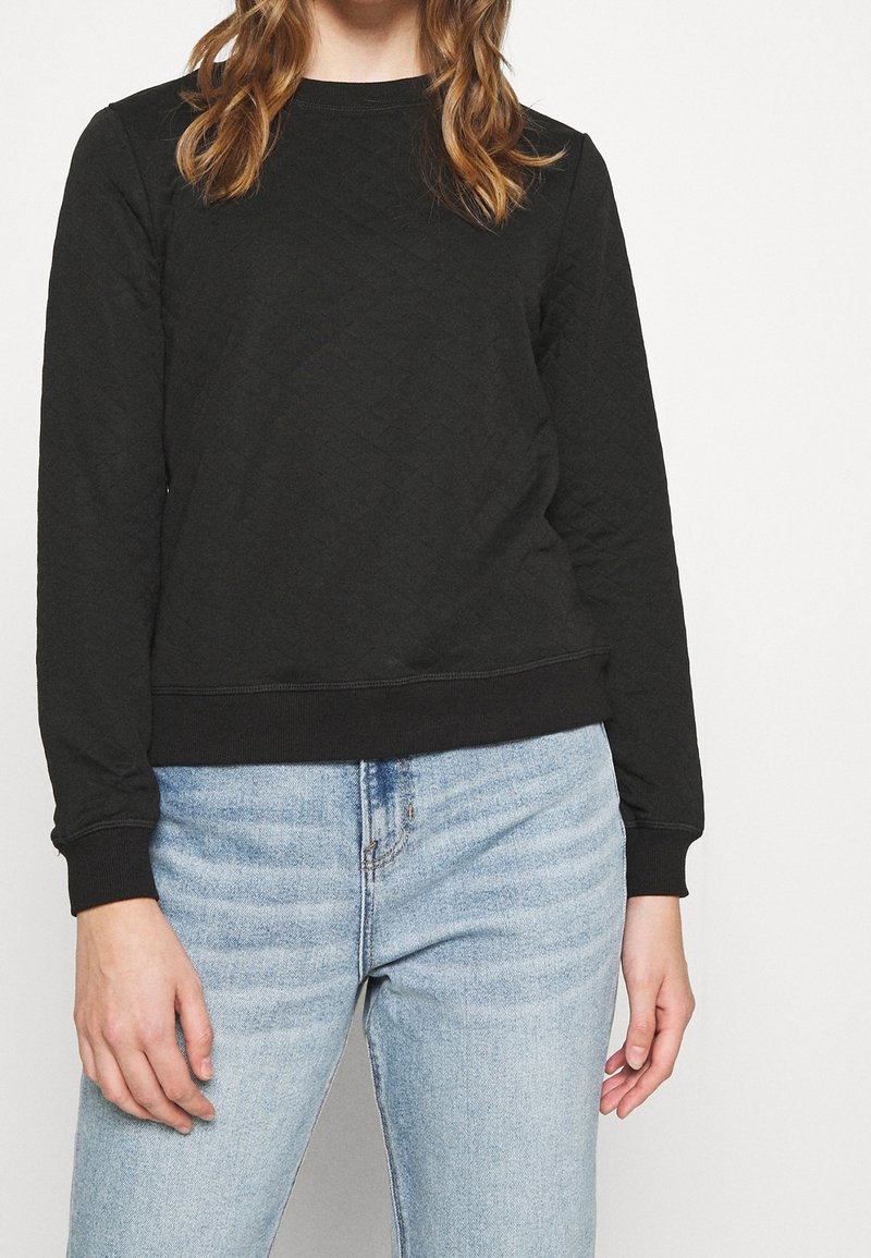 Sweatshirt noir matelassé avec un col rond, des poignets et un ourlet côtelés. Porté avec un jean en denim bleu clair, mettant en valeur un tissu texturé.