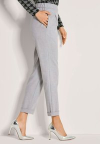 Vrouw in lichtgrijze op maat gemaakte broek met omgekrulde pijpen en zilveren hoge hakken, met hand in de zak, tegen een effen achtergrond.
