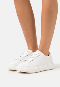 Vita låga sneakers med snörning som bärs på bar fot mot en enfärgad vit bakgrund.