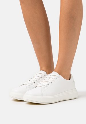 Zapatillas blancas de corte bajo con cordones, usadas sin calcetines, sobre un fondo blanco liso.