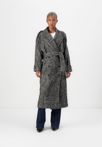 Persona con capelli biondi corti che indossa un lungo cappotto grigio a zigzag con cintura sopra jeans scuri e scarpe nere a punta, in piedi su uno sfondo bianco.