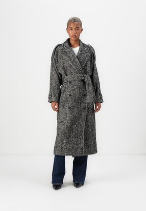 Persona con capelli biondi corti che indossa un lungo cappotto grigio a zigzag con cintura sopra jeans scuri e scarpe nere a punta, in piedi su uno sfondo bianco.