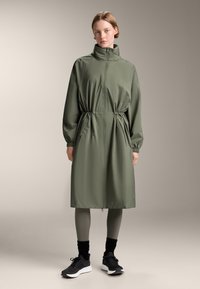 Olijfgroene, ritsjack met hoge kraag, getailleerde taille en elastische manchetten. Gstylend over grijze leggings en zwarte sportschoenen.