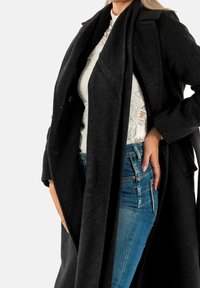 Abrigo negro de mezcla de lana con un diseño largo y abierto por delante. Presenta una tela texturizada y solapas sutiles. Lleva puesto un top de encaje blanco y jeans azules.