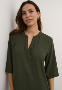 Blusa de color verde oscuro, de manga corta, con escote en V y tejido texturizado. Presenta una tapeta y un corte relajado. Se encuentra sobre un fondo claro.