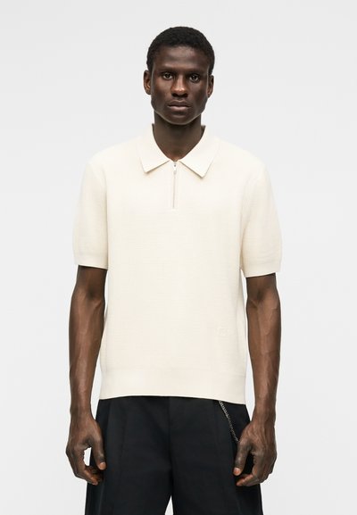 Homme portant un polo crème en maille à manches courtes avec une fermeture éclair partielle sur le devant, associé à un pantalon noir sur un fond blanc uni.