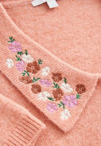 Brzoskwiniowy sweter z dzianiny z zawiniętym kołnierzem, wyszywany różowymi, białymi i brązowymi kwiatami oraz zielonymi liśćmi.