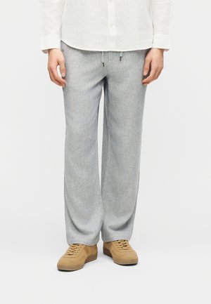 Oseba, oblečena v svetlo sive široke hlače z vrvico v pasu, belo majico z dolgimi rokavi in rjave superge z vezalkami, stoji na enobarvnem ozadju.