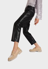 Mocassini beige in pelle con dettagli texture e suola spessa, indossati con pantaloni cropped in finta pelle nera e un top leggero viola con paillettes.