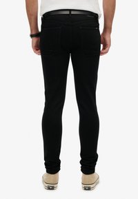 Pantalones vaqueros negros ajustados con una textura suave y lisa, que cuentan con dos bolsillos traseros y una cinturilla con un cinturón negro. Llevados con zapatillas beige.