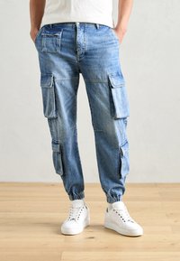 Pantalones cargo de denim azul claro con múltiples bolsillos laterales, puños elásticos en los tobillos y un corte relajado, combinados con zapatillas blancas.