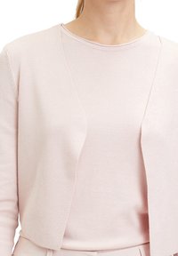 Světle růžový cardigan s hladkou texturou, otevřeným předním dílem a dlouhými rukávy, vrstvený přes odpovídající lehký top.