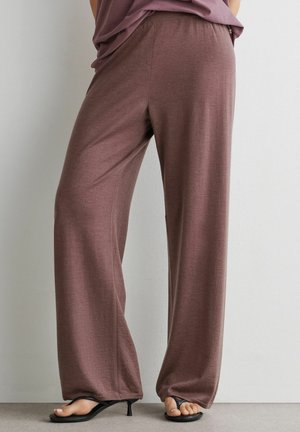 Løse brune bukser båret med sorte hælede sandaler, der viser tæerne, mod en ensfarvet lys væg og gulv baggrund.