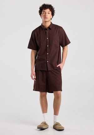 Jonge man die staat, gekleed in een bruin overhemd met korte mouwen en bijpassende korte broek, met witte sokken en beige instapschoenen.