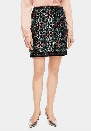 Mini-jupe noire avec des motifs circulaires verts, cuivrés et argentés. Ornements en sequins et ceinture texturée. Superposition transparente présente.