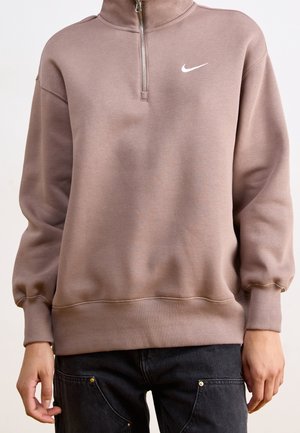 Personne portant un sweat-shirt Nike marron à fermeture éclair quart de zip avec des poignets élastiques et un pantalon noir avec des poches visibles et des boutons en laiton.