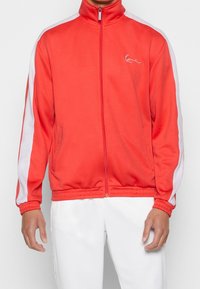 Chaqueta deportiva roja con cremallera, franjas laterales blancas, cuello alto, puños acanalados y detalle del logo en el pecho. Textura de tela suave.