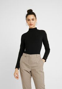Black long-sleeve top with a high neck and lace accents on the sleeves, paired with light brown plaid trousers. Smooth texture.