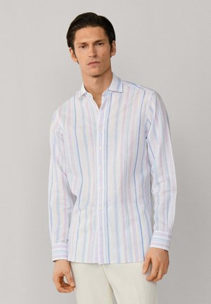 Homme portant une chemise blanche à manches longues et boutonnée avec des rayures pastel verticales, debout devant un fond gris clair.