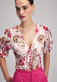 Bluse mit floralem Print in Hellrosa, mit elastischer Raffung, tiefem V-Ausschnitt und Bindeband, kombiniert mit hochgeschnittenen rosa Hosen.