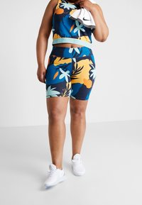 Květinový sportovní set zahrnující bezrukávový crop top a šortky s vysokým pasem v námořnické modři s oranžovými, žlutými a světle modrými vzory; doplněné bílými teniskami.