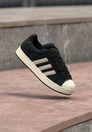 Zapatilla de gamuza negra con tres franjas blancas, puntera redonda y cordones negros, con superficie texturizada y entresuela crema.