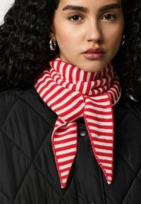 Écharpe rayée rouge et blanche en matière tricotée, nouée autour du cou, placée sur une veste matelassée noire. Détails texturés visibles.