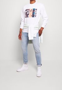 Camisa blanca de manga larga con un gráfico impreso en cuatro paneles. Jeans azul claro, sudadera verde menta atada a la cintura y zapatillas blancas.