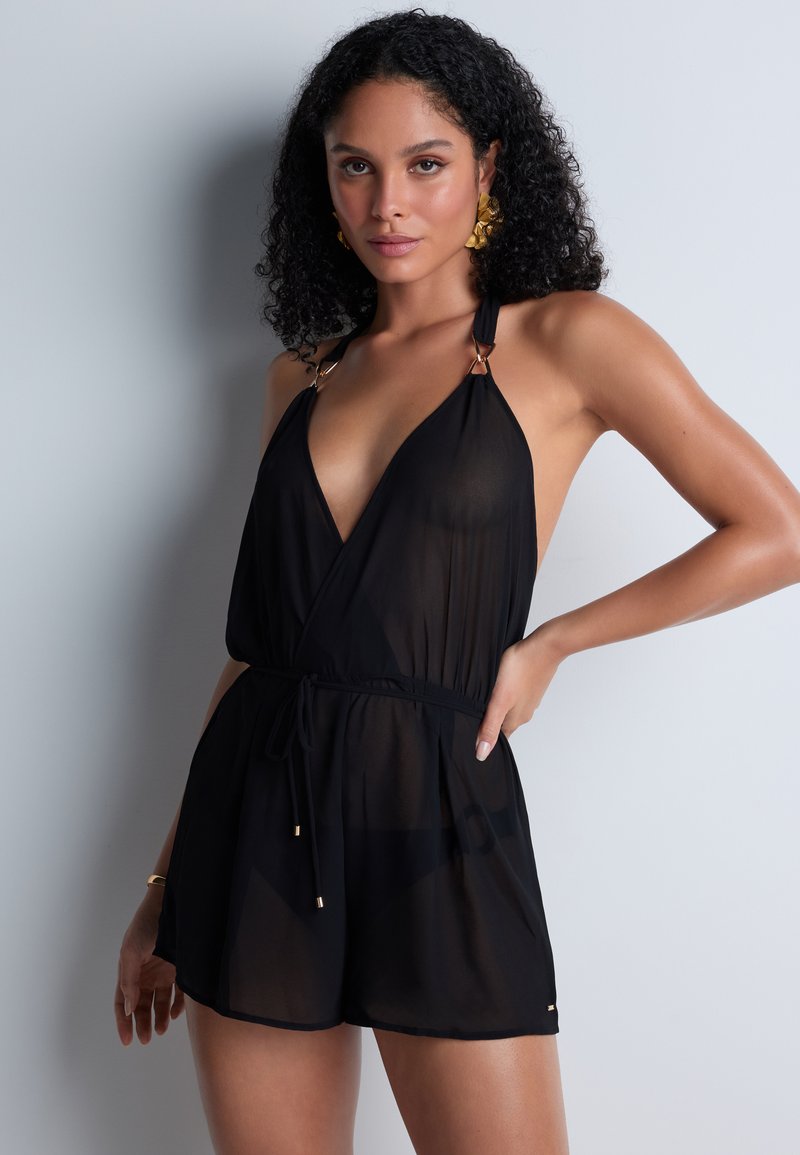 Combinaison noire sans manches en tissu transparent, avec un décolleté en V profond, des bretelles réglables avec des accessoires en or, et une taille cintrée avec un lien.