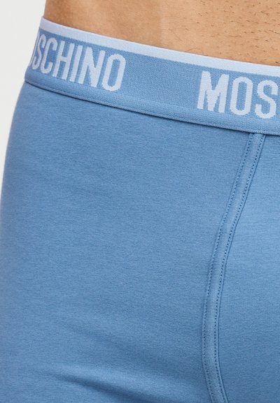Κοντινό πλάνο στη ζώνη και το ύφασμα γαλάζιου αντρικού εσώρουχου, που δείχνει μερικό λευκό λογότυπο "MOSCHINO" στη ελαστική ταινία.