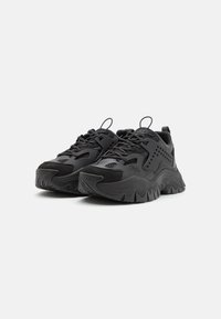 Baskets de sport noires en matériaux synthétiques avec une surface texturée, semelle épaisse et système de laçage bien visible. Comprend des accents en maille.