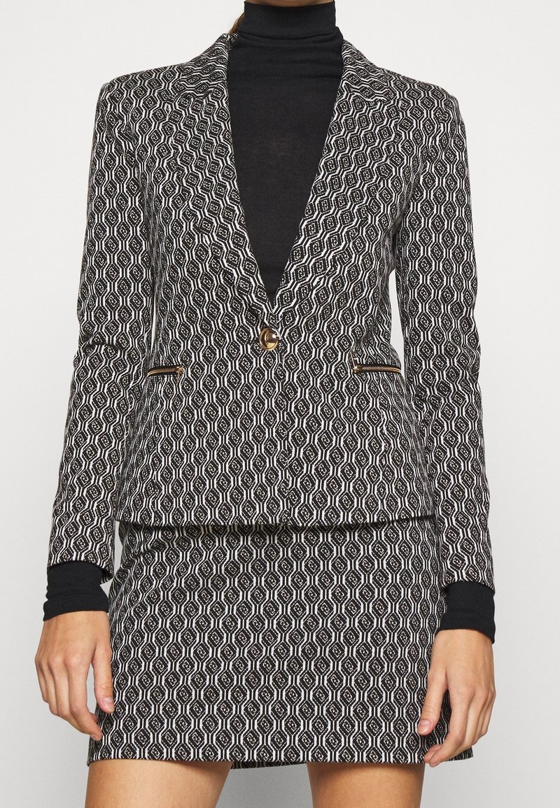 Femme portant un blazer à motif géométrique noir et blanc et une jupe assortie par-dessus un top à col roulé noir.
