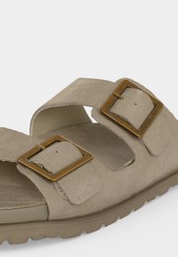 Μπεζ σανδάλι με δύο ρυθμιζόμενους ιμάντες που διαθέτουν μεγάλες ορθογώνιες χάλκινες αγκράφες και μαξιλαρωμένη σόλα με υφή για καλύτερο κράτημα.