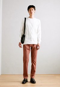 Valkoinen vaaleanpunaisella neuleella, ruskeat housut, musta olkalaukku, mustat loaferit. Seisova asento vaaleaa taustaa vasten.