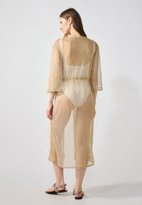 Kimono de longitud media en tono dorado, de tela transparente, con mangas de tres cuartos y una cinturilla ajustable, que presenta un acabado texturizado y aberturas laterales.