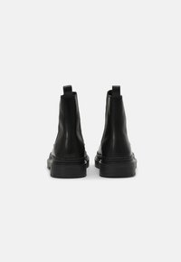 Botas negras hasta el tobillo hechas de cuero liso, con una lengüeta, punta redondeada y suela de goma texturizada. Diseño simple sin patrones visibles.