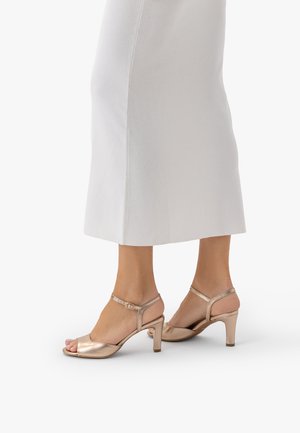 Stopy w metalicznych sandałach na obcasie w kolorze różowego złota z paskiem wokół kostki, zestawione z białą spódnicą midi na jednolitym białym tle.