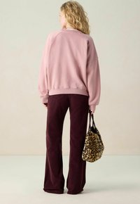 Femme aux cheveux blonds ondulés portant un sweat à capuche rose ample et un pantalon bordeaux, tenant un sac à main en fausse fourrure à imprimé léopard, de dos.