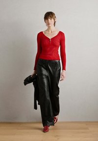 Haut rouge à manches longues côtelé avec un détail zippé, associé à un pantalon en cuir noir taille haute à jambes larges et des chaussures rouges, le tout sur un fond neutre.