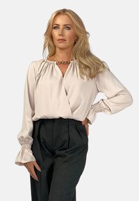 Jasnoróżowa bluzka z zebranym dekoltem i akcentem w postaci łańcuszka, z długimi rękawami zakończonymi marszczonymi mankietami, noszona z wysokimi spodniami w paski.