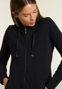 Felpa nera con zip, caratterizzata da un pannello inferiore testurizzato, lacci in satin e dettagli in metallo sulla zip. Tessuto morbido con maniche lunghe e vestibilità aderente.