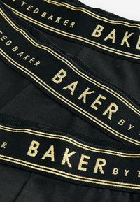 Zwarte tailleband met gouden opdruk "TED BAKER" en "BAKER by Ted Baker." De stof lijkt zacht en rekbaar met een gladde textuur.