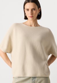 Beige strikket genser med avslappet passform, korte ermer og bred halsåpning. Glatt tekstur, minimale sømmer og et uformelt design.