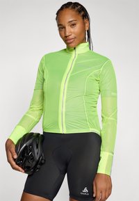 Giacca da ciclismo leggera, trasparente e verde neon con maniche lunghe e zip frontale, abbinata a pantaloni da ciclismo neri imbottiti. Casco tenuto in mano.
