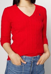 Pull rouge en maille torsadée avec un col en V et des manches trois-quarts, présentant un motif texturé. Associé à un short en jean taille haute.