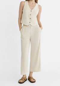 Beige Leinenweste mit V-Ausschnitt und fünf Knöpfen, kombiniert mit beige Hochbund-Hosen mit weitem Bein. Braune flache Sandalen runden den Look ab.