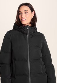 Woman with short dark hair wearing a black padded winter jacket with hood, zipped up, looking slightly to the side and smiling.